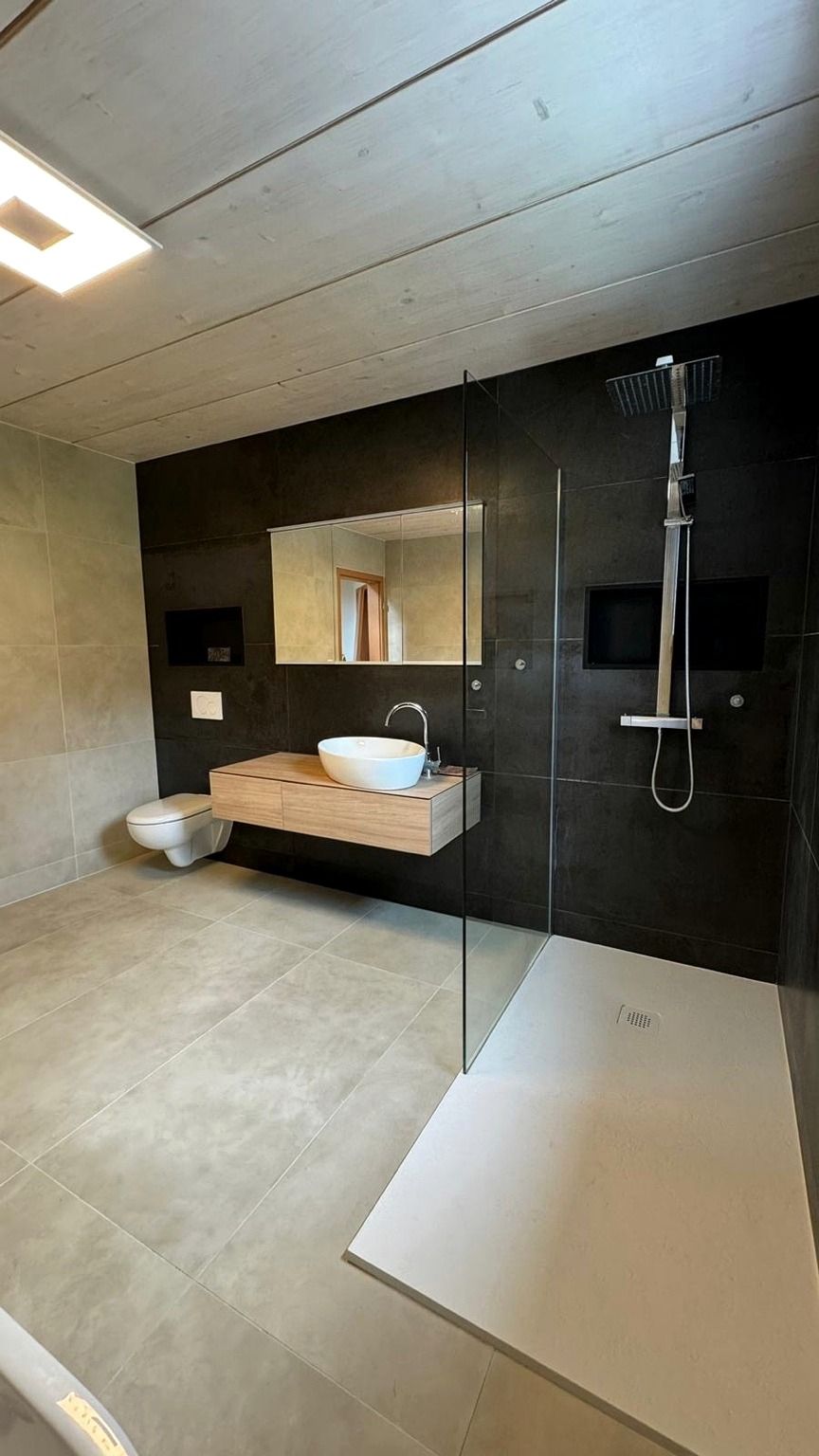 Salle de bain rénovée à Belmont-sur-Lausanne, vue d’ensemble après travaux, avec murs en grands formats foncés, meuble vasque suspendu en bois, WC suspendu, douche de plain-pied vitrée et sol clair