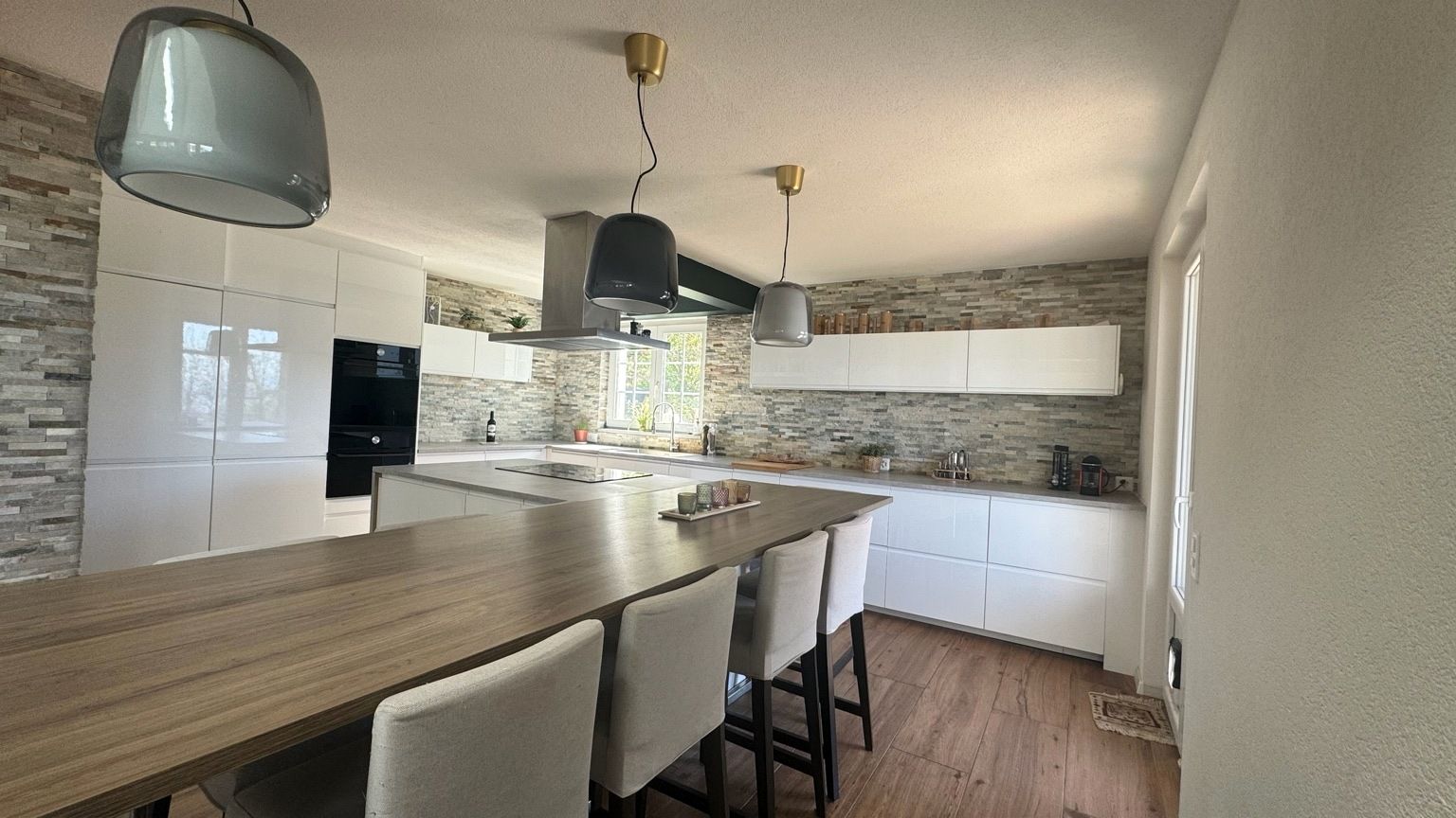 Cuisine rénovée à Épalinges, avec grande table de repas effet bois et tabourets blancs, îlot, façades blanches brillantes et parement en pierre naturelle