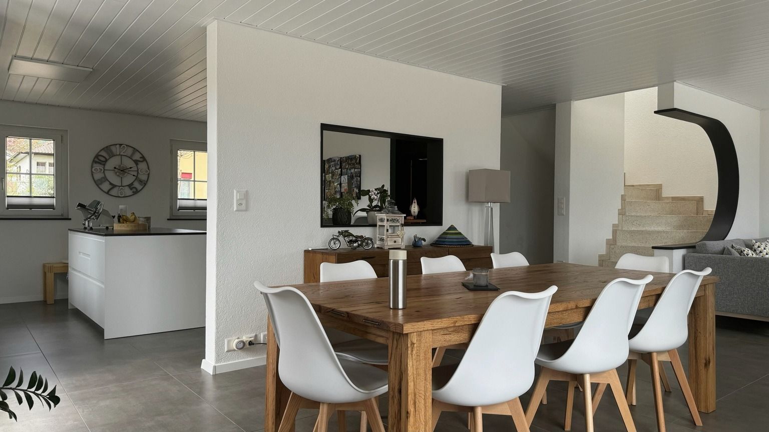 Espace de vie rénové à Bevaix, avec table en bois massif et chaises blanches, cuisine ouverte, escalier et sol en grand format gris
