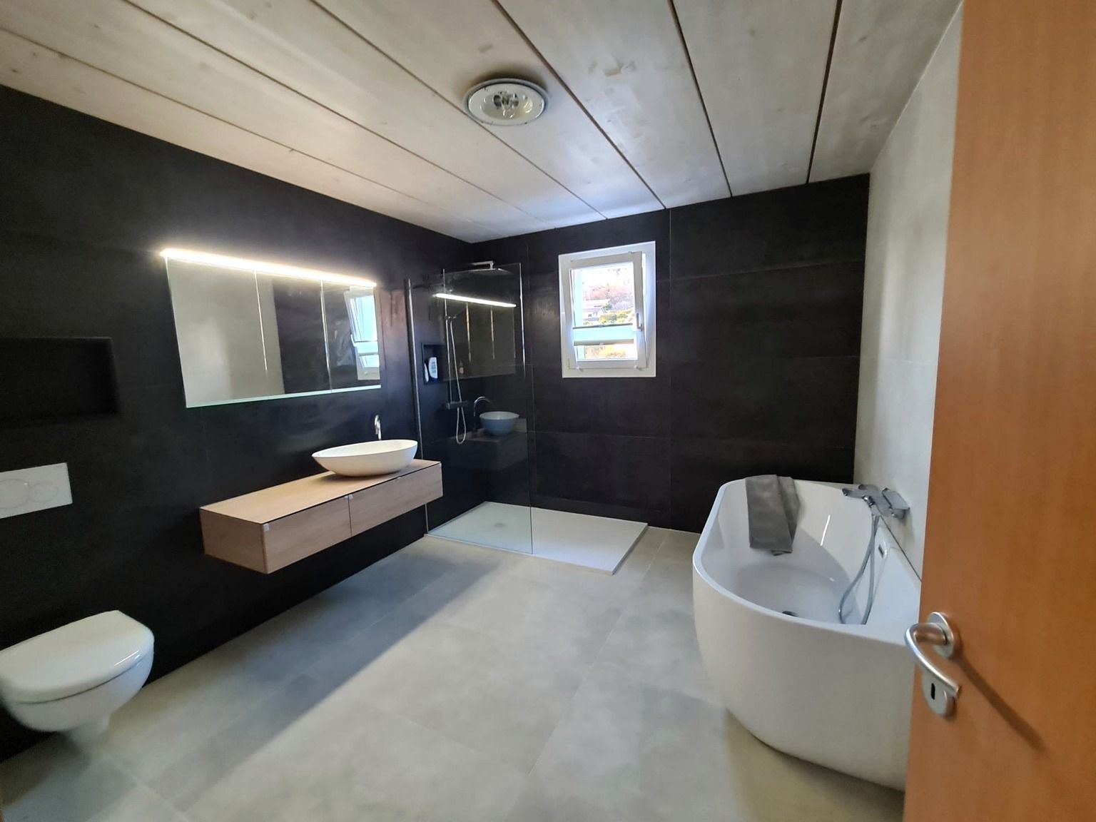 Vue générale de la salle de bain après rénovation, avec meuble vasque suspendu et miroir éclairé, douche de plain-pied, baignoire îlot et sol en grand format clair