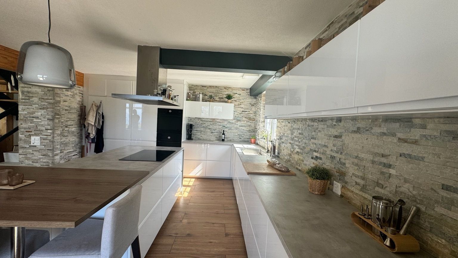 Seconde vue de la cuisine, avec îlot et plan de cuisson, long plan de travail effet béton, armoires hautes blanches et crédence en parement de pierre
