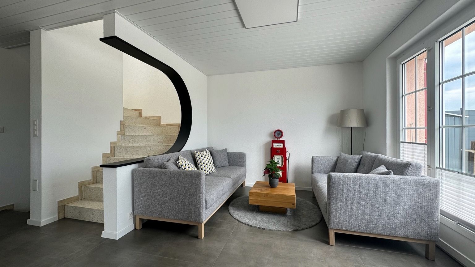 Séjour lumineux avec deux canapés gris, table basse en bois et escalier sous une courbe sombre, devant de grandes fenêtres
