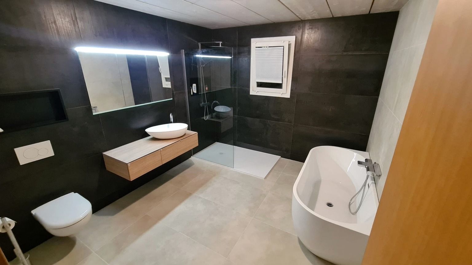 Autre vue de la salle de bain rénovée, avec la baignoire îlot au premier plan, le meuble vasque suspendu et la douche de plain-pied au fond