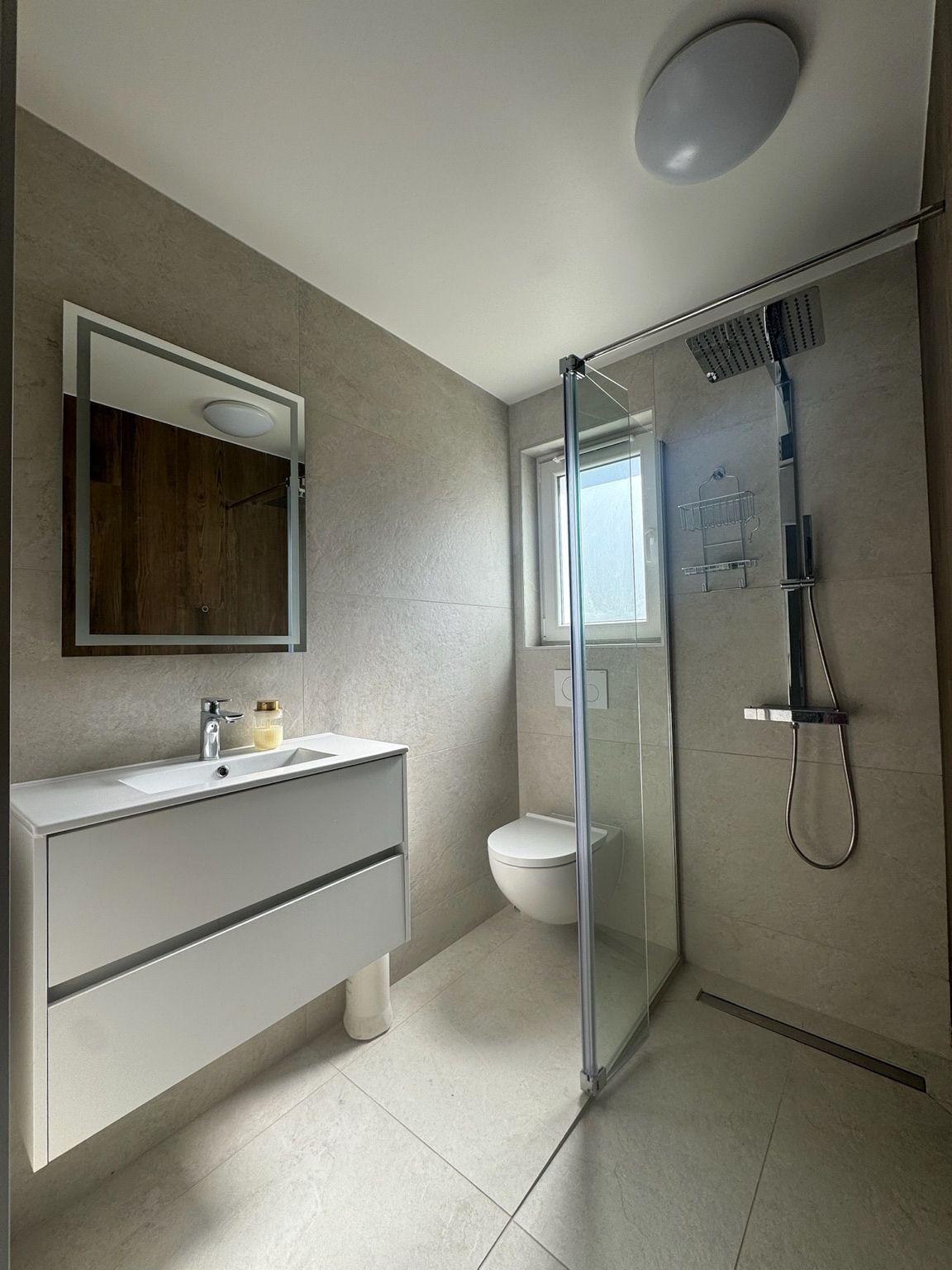 Salle de bain rénovée, avec meuble vasque blanc suspendu, miroir éclairé, WC suspendu et douche de plain-pied vitrée en grands formats clairs