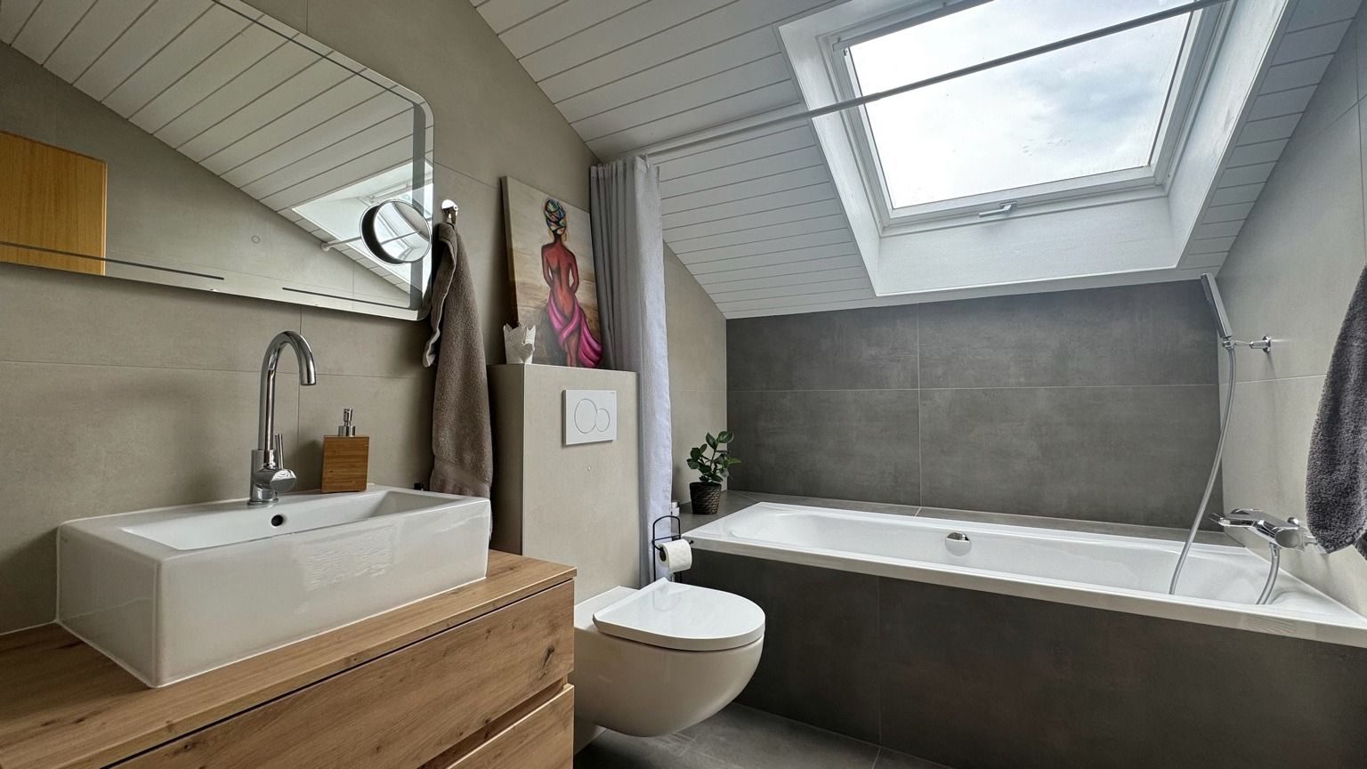 Salle de bain sous toiture, avec meuble vasque en bois, WC suspendu, baignoire habillée de grands carreaux gris foncé et fenêtre de toit
