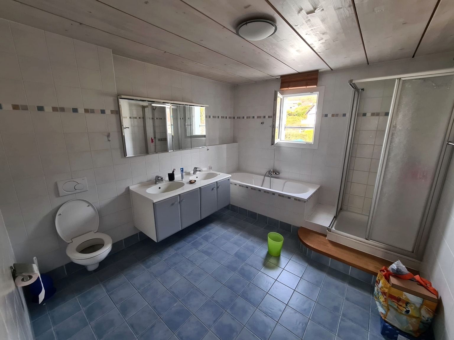 La salle de bain avant rénovation, avec carrelage blanc et frise, sol en carrelage bleu, double lavabo, baignoire encastrée et cabine de douche
