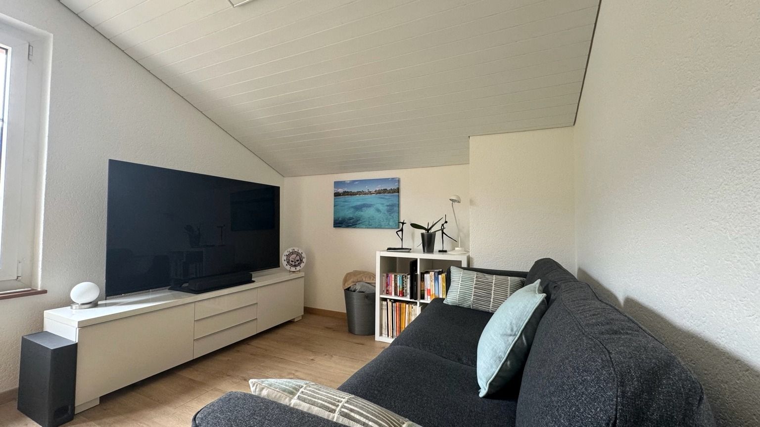 Petit séjour sous toiture, avec canapé gris foncé, meuble TV blanc et sol en parquet clair
