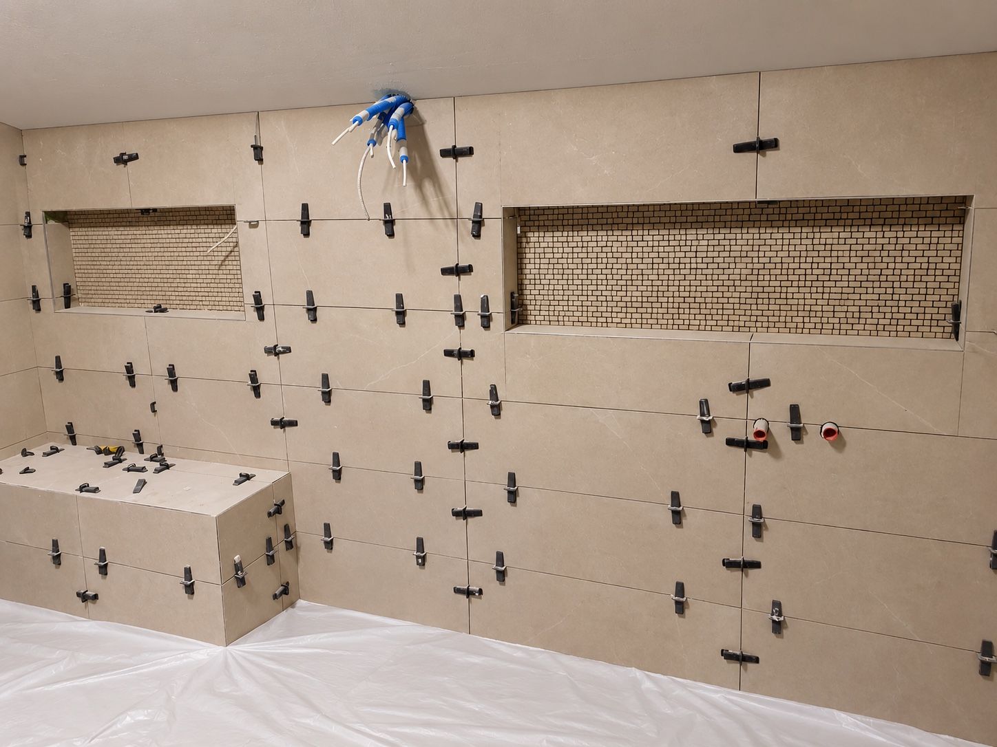 Chantier de salle de bain en cours : grands carreaux posés avec leurs croisillons de nivellement, niches murales en mosaïque, banc carrelé et sol protégé par une bâche blanche